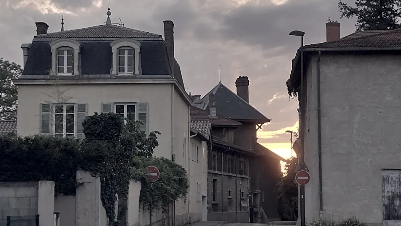 Haut d’une rue de Charbonnières-les-Bains au soleil couchant, entre une maison bourgeoise à lucarnes et des bâtiments anciens aux toits pentus.