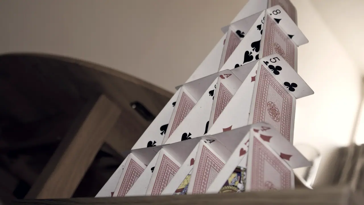 Château de cartes à jouer dressé sur plusieurs étages, les cartes appuyées deux à deux sous des cartes posées à plat, devant un mur clair.