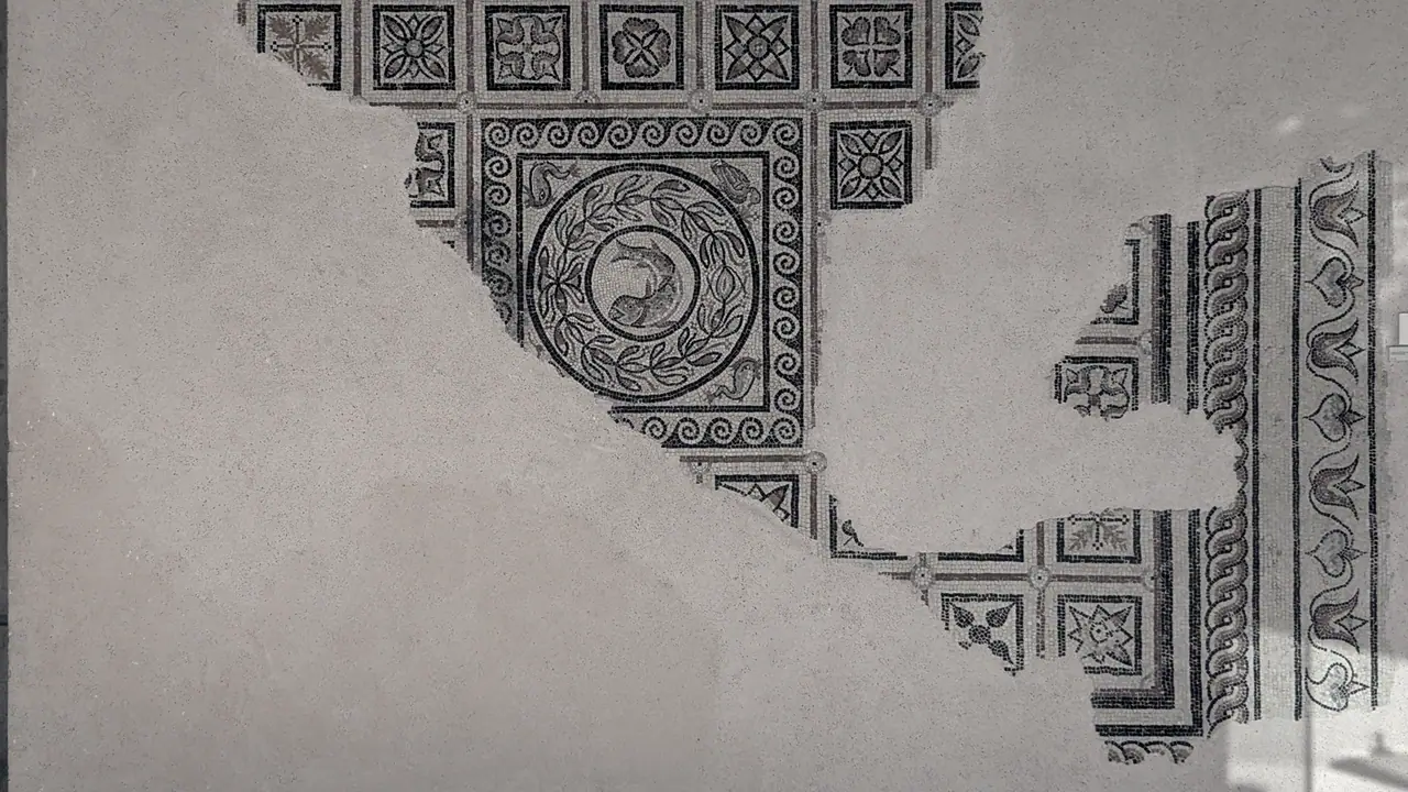 Fragment de mosaïque romaine fixé au mur, avec un médaillon au poisson et des carrés à rosaces qui s'interrompent sur de larges vides de mortier nu.