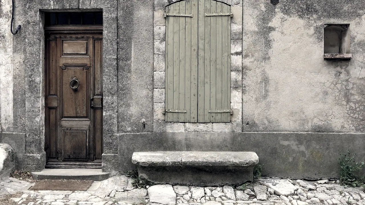 Façade ancienne d'une maison de village, avec une porte de bois à panneaux et son heurtoir, une fenêtre aux volets clos, une niche dans le mur et un banc de pierre sur le pavé.