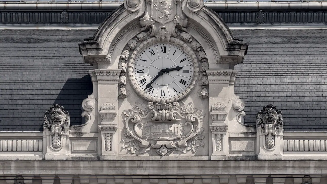 Grande horloge à chiffres romains encastrée dans un fronton de pierre sculpté, entre deux mascarons de lion, devant un toit d'ardoise.