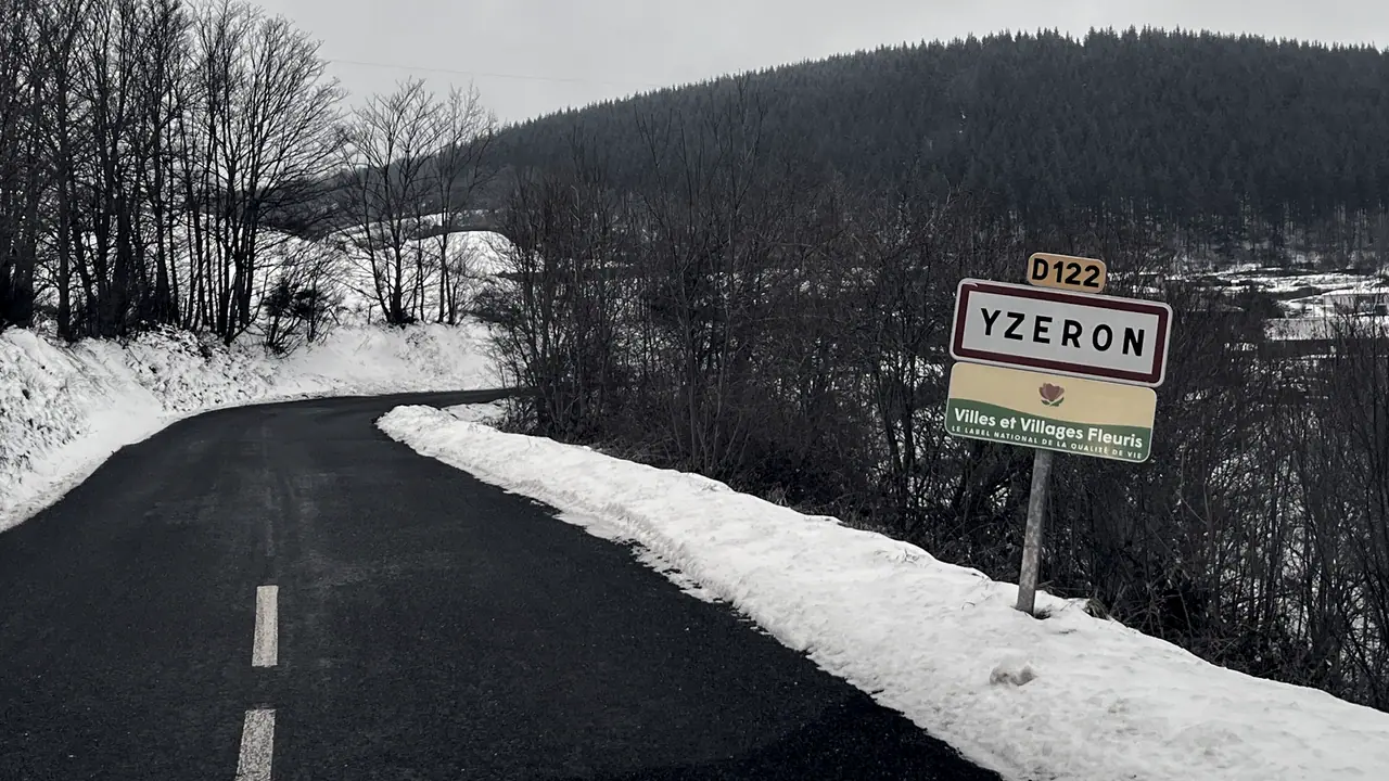Panneau d'entrée de la commune d'Yzeron au bord d'une route de montagne bordée de neige, devant une colline couverte de sapins.