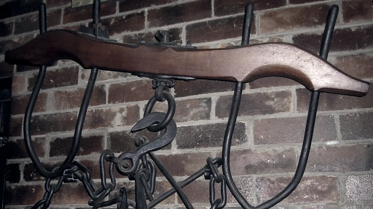 Joug de bœufs en bois accroché contre un mur de briques, avec ses deux arceaux de fer et la chaîne à crochet qui pend en son milieu.