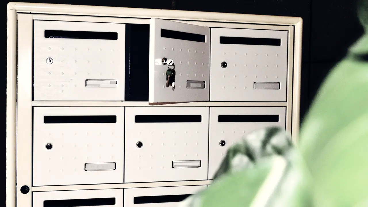 Bloc de boîtes aux lettres à plusieurs cases, une porte entrouverte avec son trousseau de clés resté dans la serrure, et un feuillage flou qui entre dans le cadre.