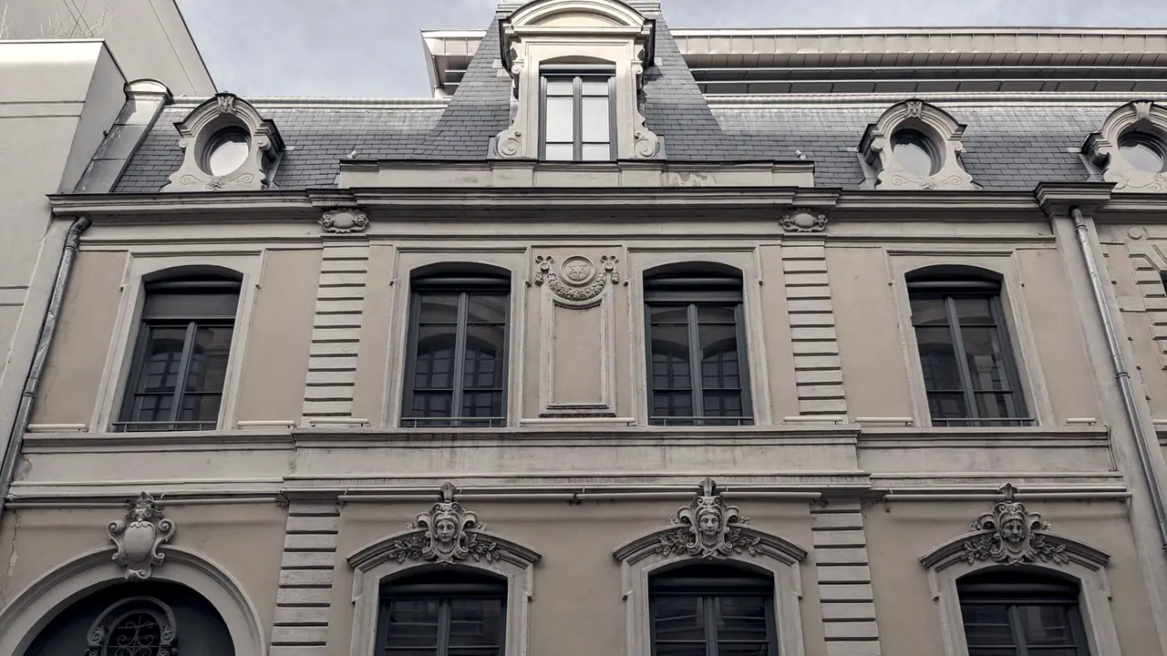 Façade d'immeuble ancien prise depuis la rue, avec un toit d'ardoise à lucarnes, des corniches moulurées et des mascarons sculptés au-dessus des fenêtres.