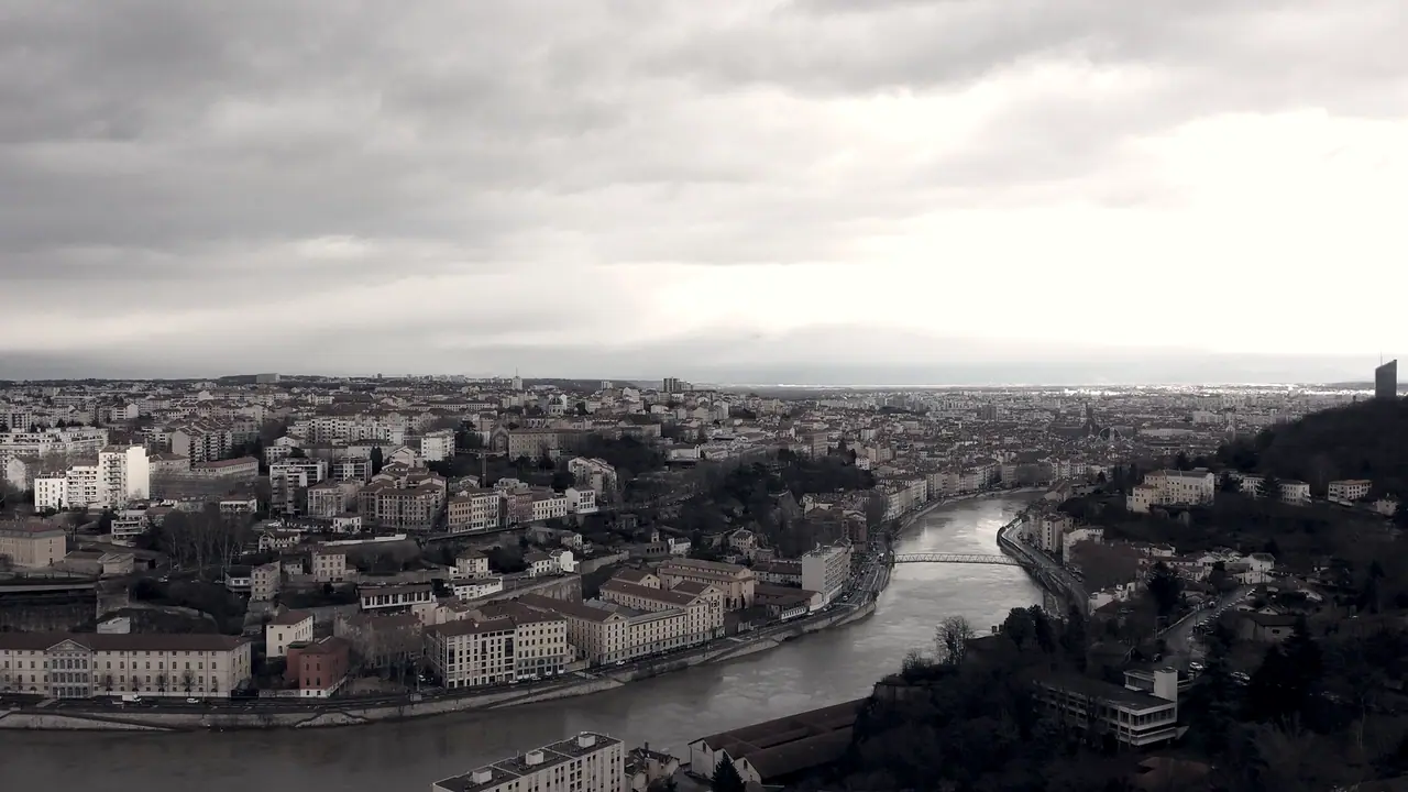 Vue plongeante sur une ville traversée par une rivière, avec ses toits serrés, sa passerelle et les collines jusqu'à l'horizon.