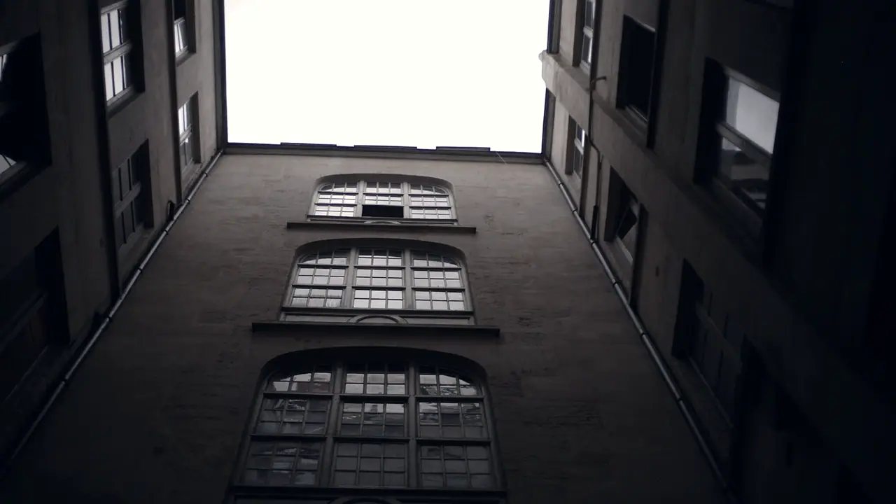 Cour étroite photographiée vers le ciel, avec les trois hautes fenêtres superposées d'une cage d'escalier sur le mur qui la ferme.