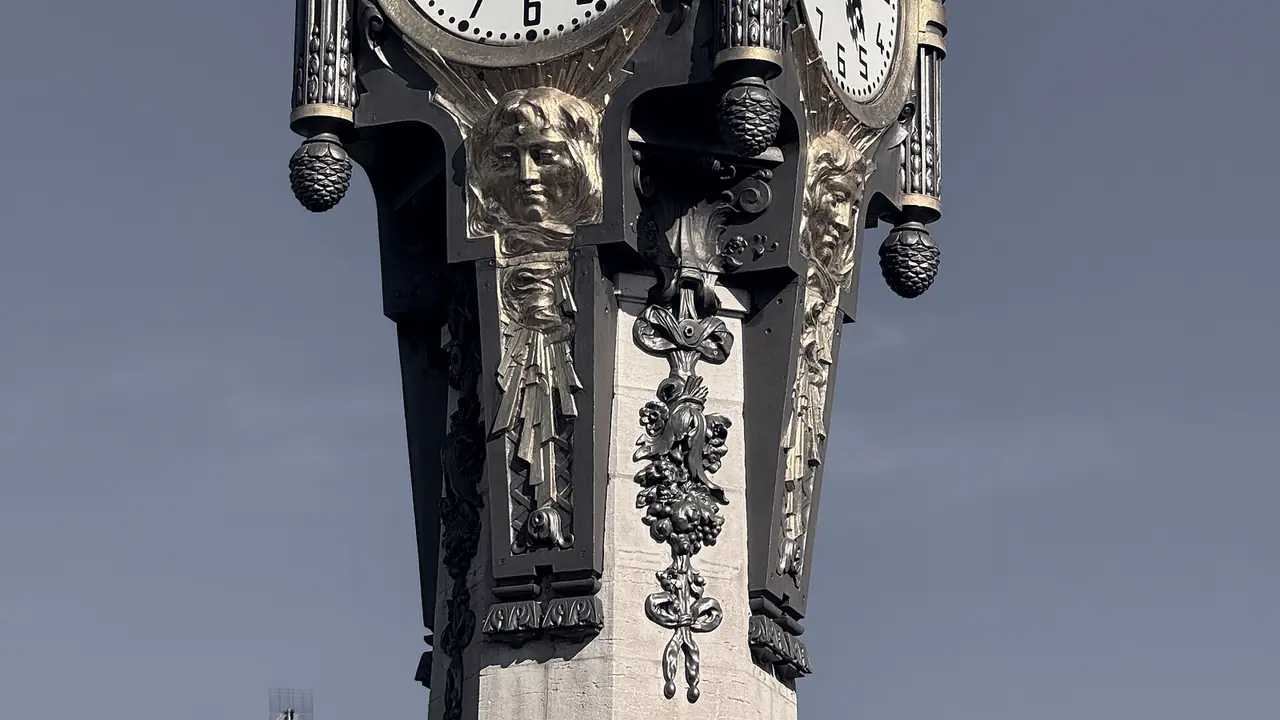 Pilier d'une horloge monumentale, avec des mascarons sculptés, des guirlandes de fruits et des pommes de pin en fonte sous ses cadrans.