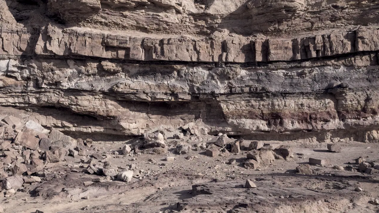 Paroi de roche sédimentaire : des couches d'épaisseurs et de teintes différentes se superposent en bandes horizontales, au-dessus d'un sol jonché d'éboulis.