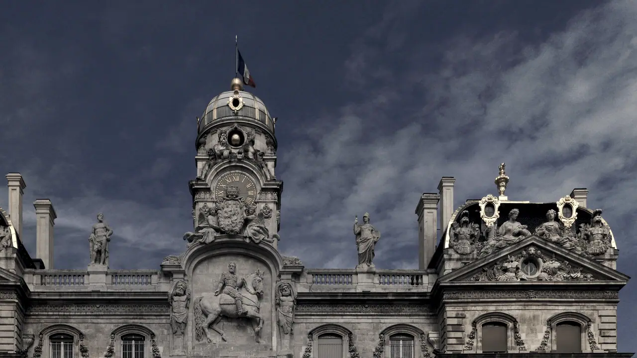Couronnement d'un hôtel de ville avec son dôme, des statues alignées sur la balustrade et un relief équestre sous un ciel chargé de nuages.