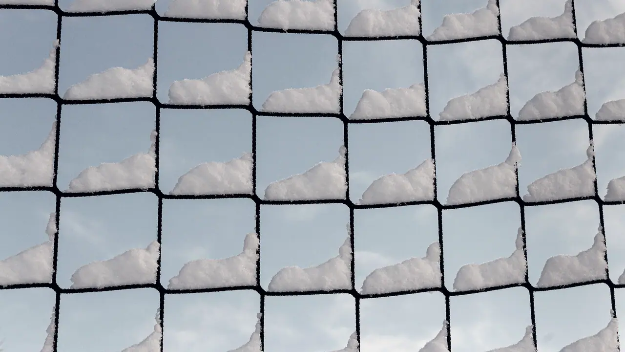Mailles carrées d'un filet de protection sportif, chacune portant un petit bourrelet de neige, devant un ciel à peine voilé.
