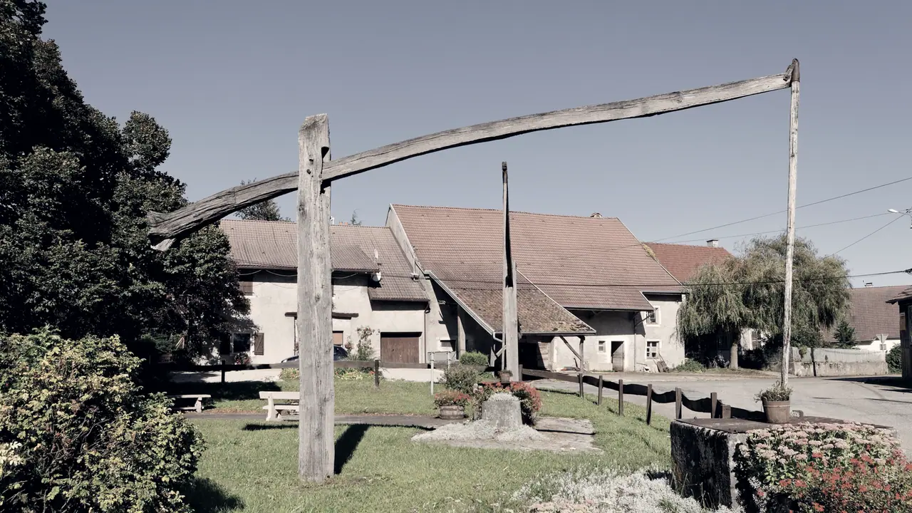 Puits à balancier dans un village : une longue perche de bois pivote au sommet d'un poteau, son gros bout faisant contrepoids, l'autre retenant la tige du seau.