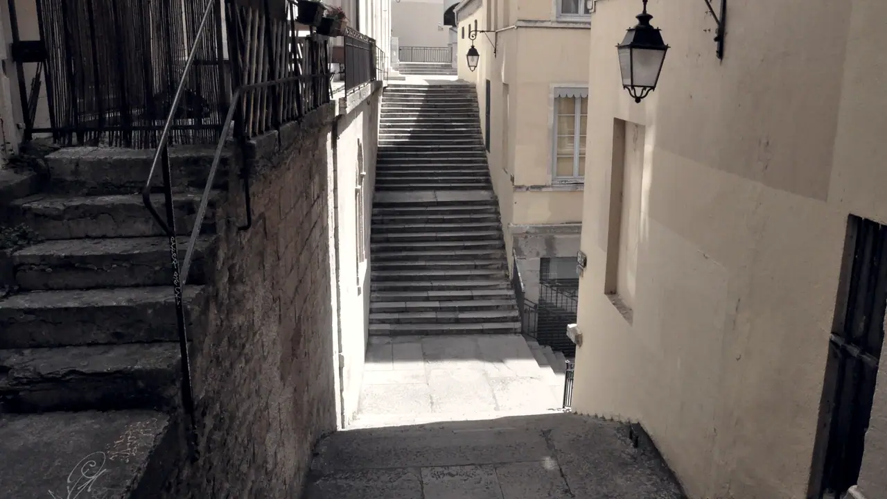 Ruelle étroite entre deux murs, avec un escalier de pierre qui monte vers le fond et une lanterne fixée en applique sur la façade.