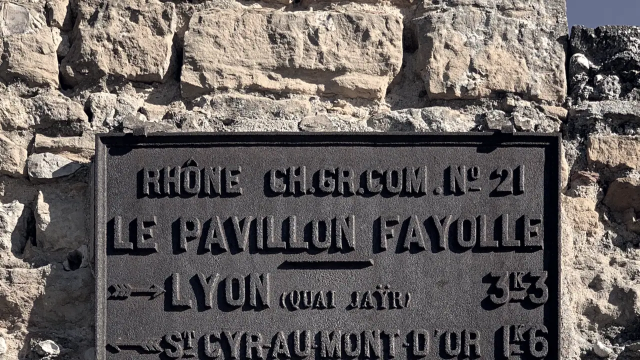 Plaque de fonte aux lettres en relief, vissée sur un mur de pierres appareillées, indiquant des directions et des distances vers Lyon.
