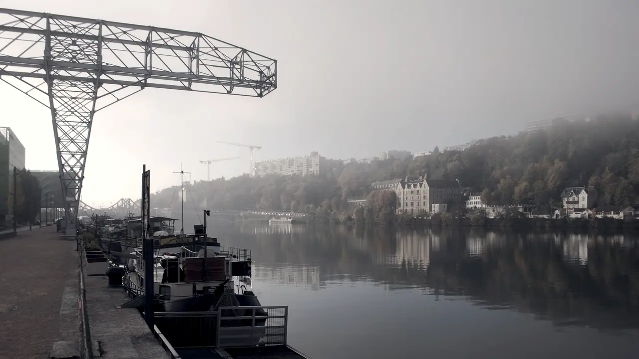 Péniches amarrées le long d'un quai dans la brume, sous une ancienne grue métallique, avec un coteau boisé et des maisons sur l'autre rive.