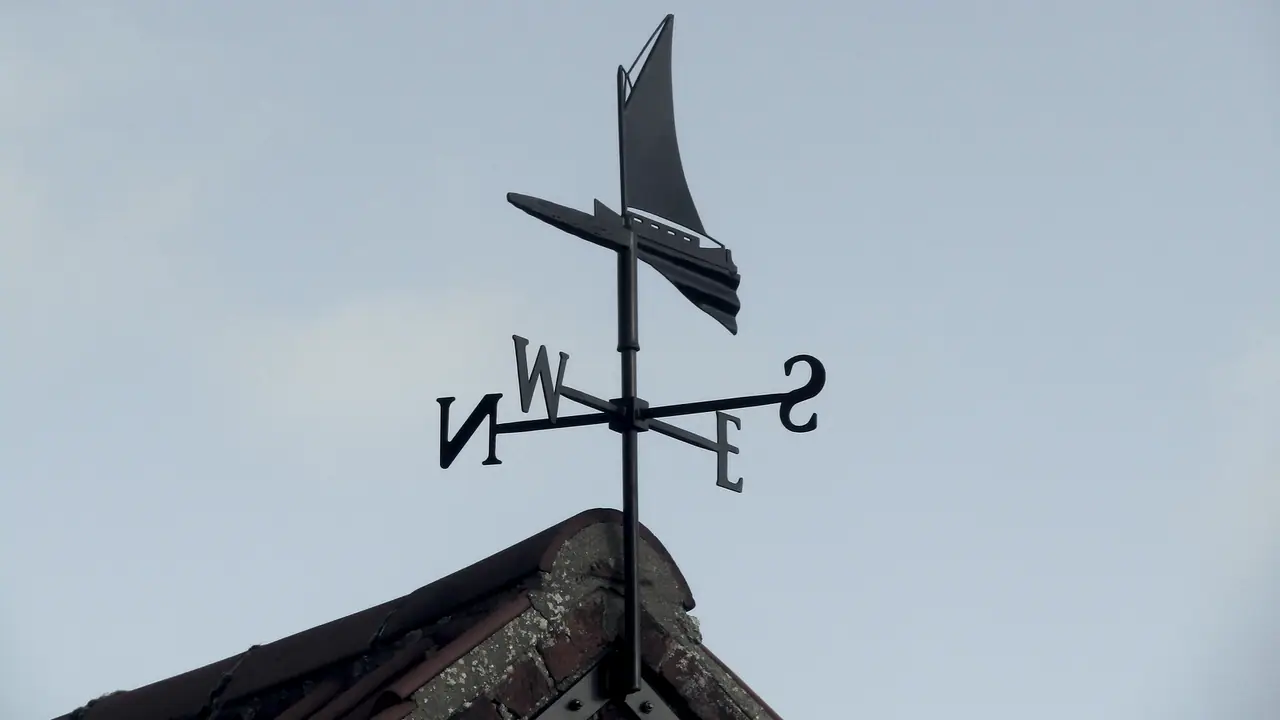 Girouette de métal en forme de bateau à voile, dressée sur le faîtage d'un toit de tuiles : sous la voile, les lettres des quatre points cardinaux se croisent.