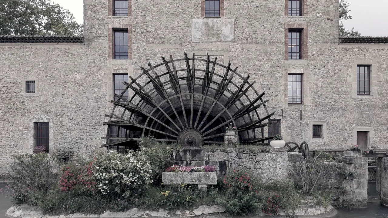 Grande roue à aubes de bois dressée contre la façade de pierre d'un ancien moulin, au-dessus d'un massif fleuri planté au bord de l'eau.