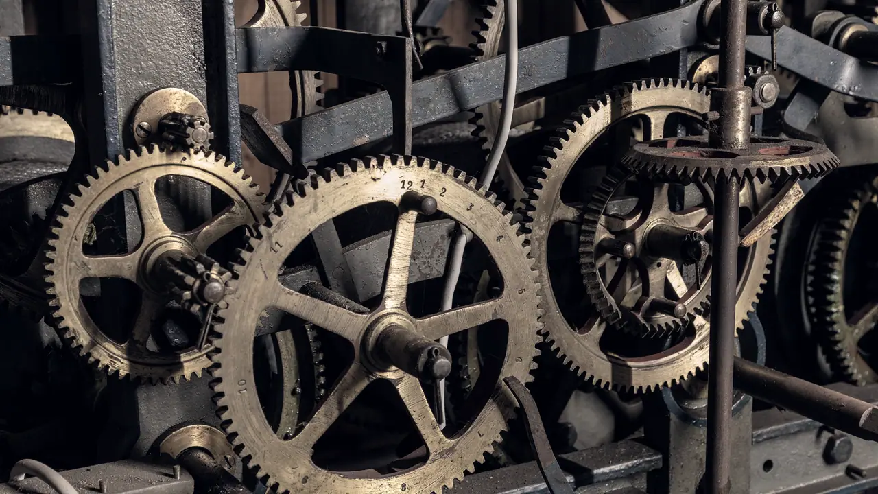 Rouages de laiton du mécanisme d'une ancienne horloge d'église : plusieurs roues dentées s'engrènent autour d'un axe vertical, sur un bâti de métal sombre.