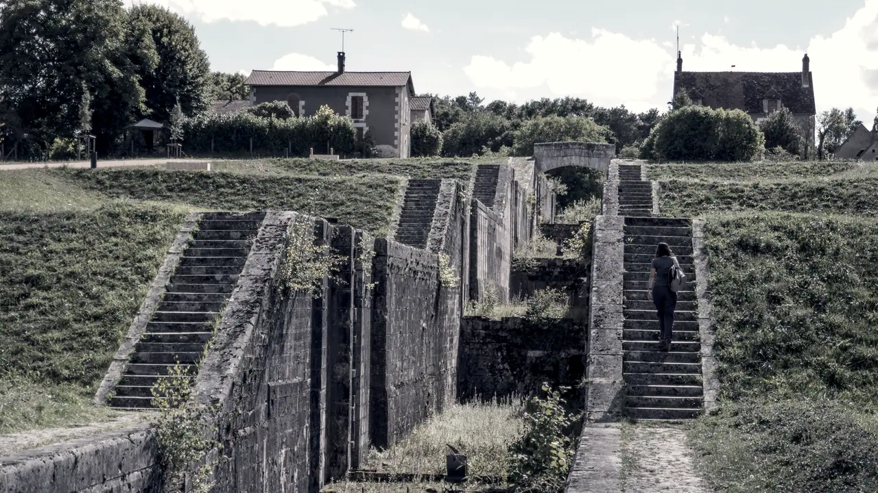 Échelle d'écluses désaffectée à Rogny : des sas de pierre se succèdent en montant le long d'un coteau herbeux, flanqués d'escaliers, avec une promeneuse de dos sur les marches.