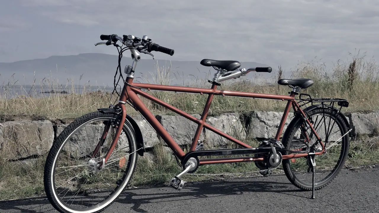 Tandem garé sur un chemin goudronné devant un muret de pierres, avec ses deux selles et ses deux guidons sur un seul cadre, et une montagne au loin.