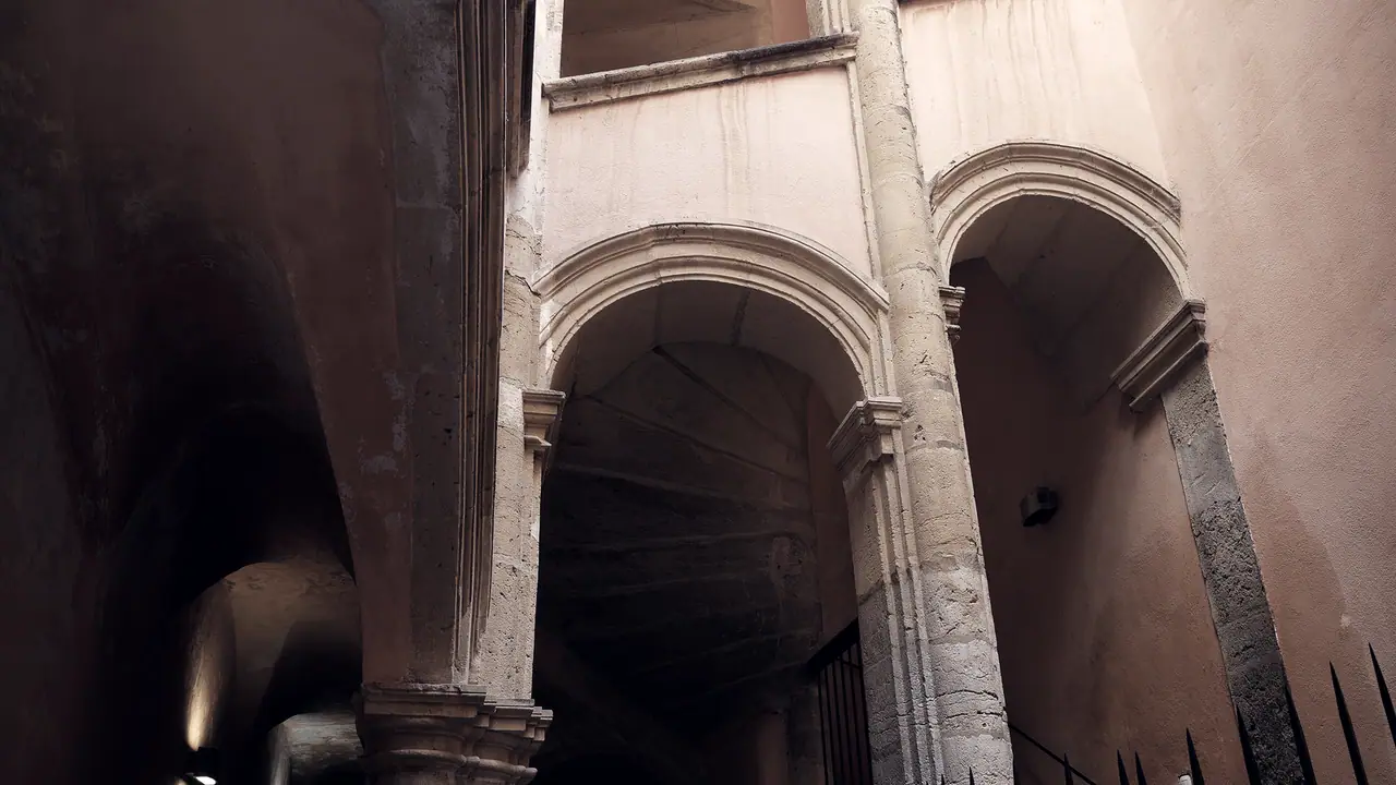 Arcades en pierre d'une cour intérieure, avec des piliers massifs et un escalier sombre derrière la grille du fond.