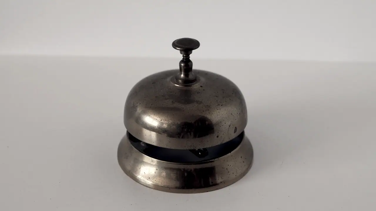 Sonnette de comptoir en métal posée seule sur une table nue devant un mur uni, son bouton dressé au sommet du timbre.