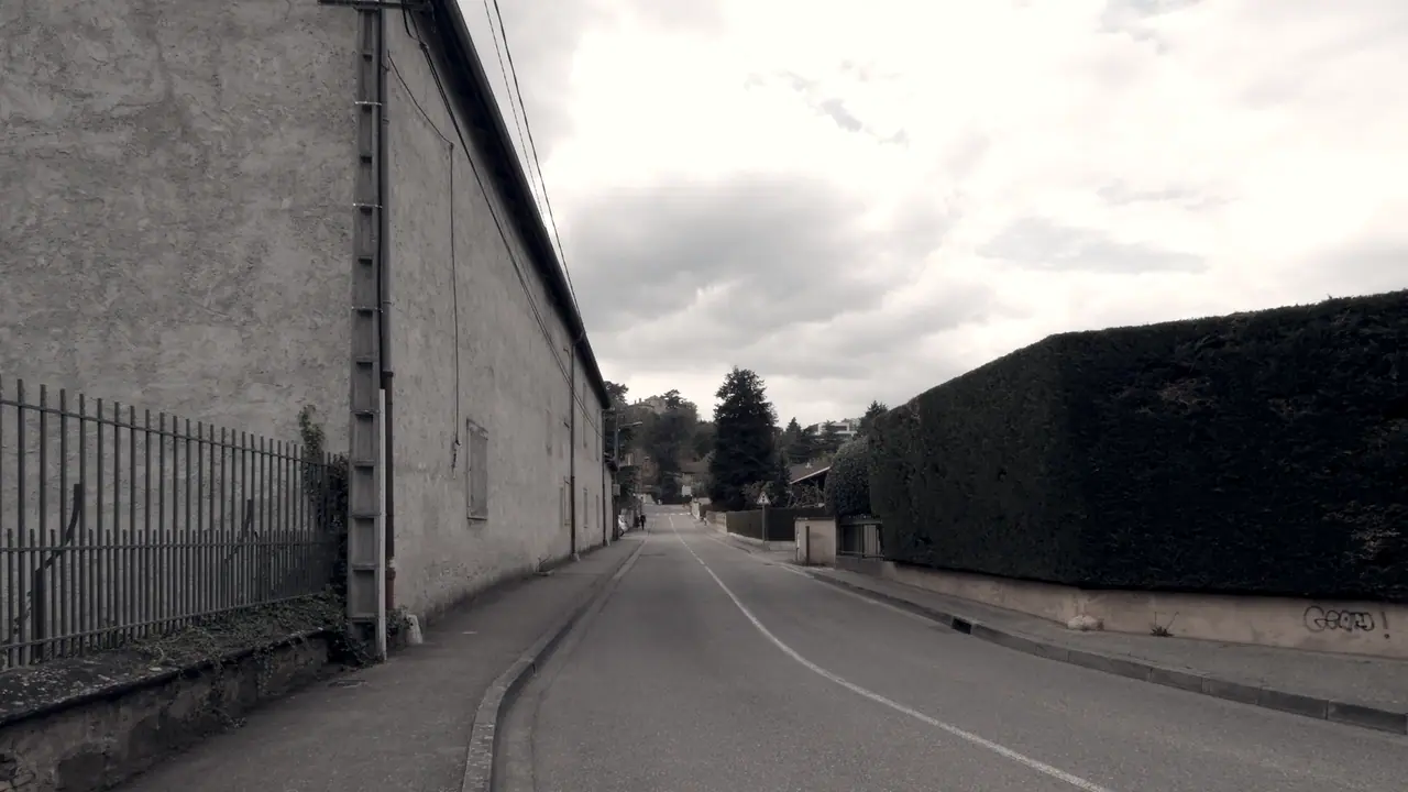 Route déserte entre un long mur crépi bordé d'une grille et une haie taillée très haute sur le côté opposé.