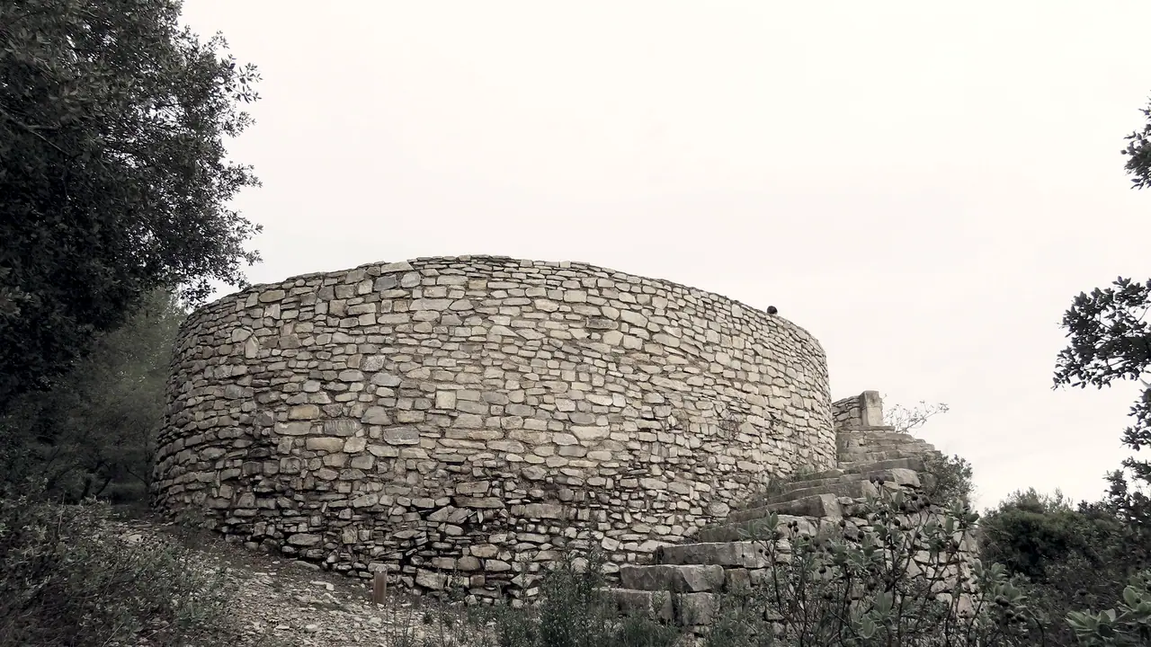 Tour ronde en pierres sèches dans la garrigue, avec un escalier de dalles qui monte le long de son flanc jusqu'au sommet.