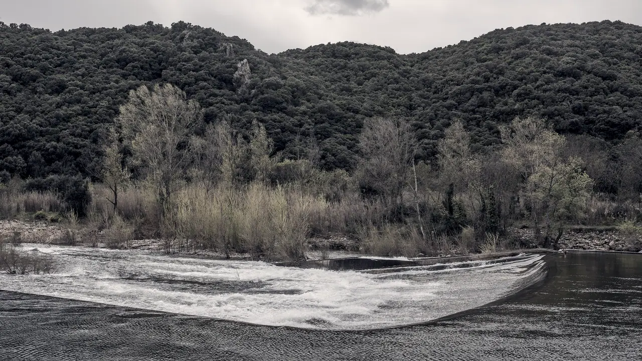 Rivière qui déborde en nappe blanche par-dessus un seuil de pierre courbe, au pied d'une colline couverte de chênes verts.