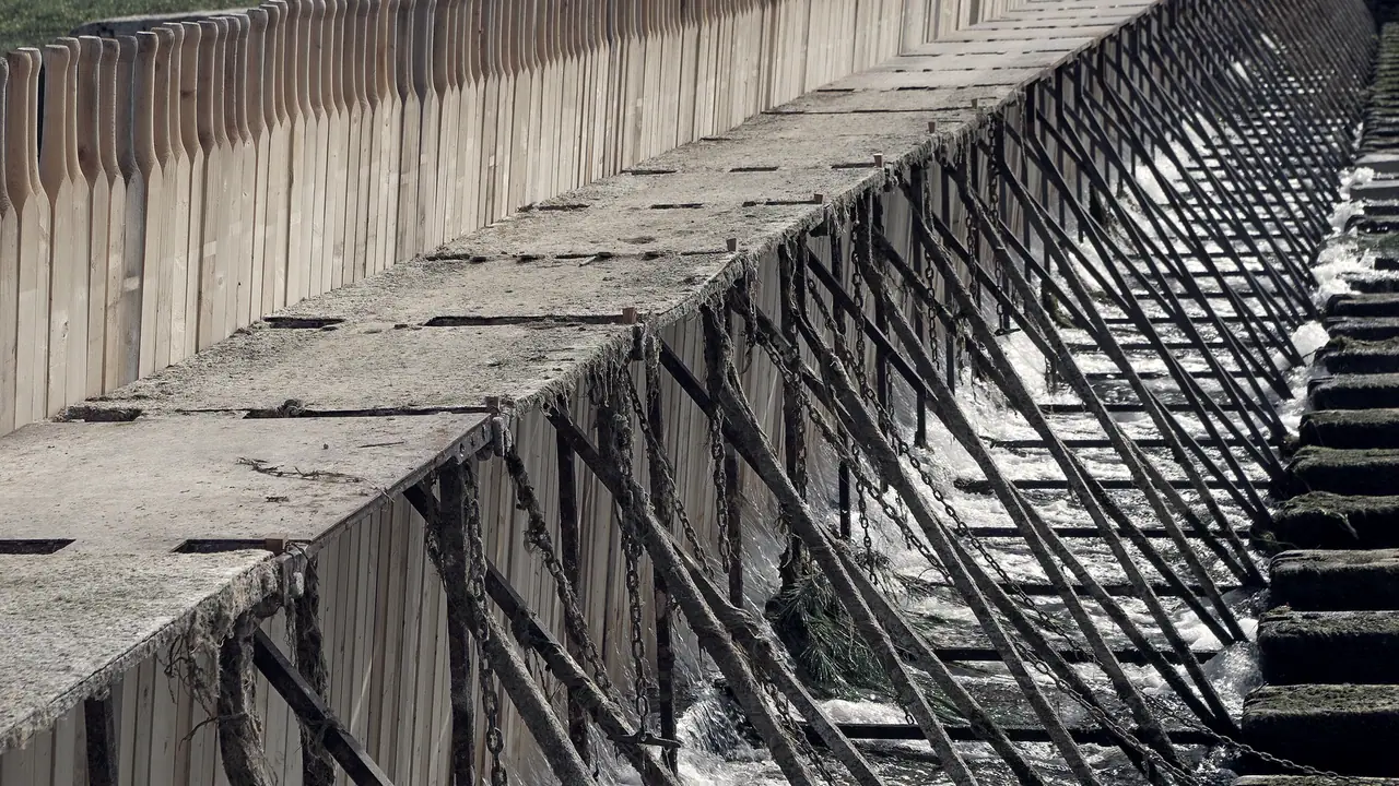 Barrage à aiguilles sur une rivière : une longue rangée de perches de bois appuyées en biais contre une passerelle de béton.