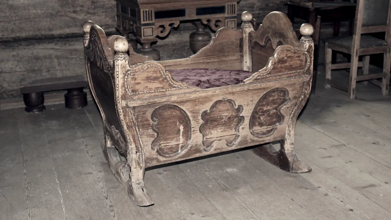 Berceau ancien en bois sculpté monté sur des patins courbes, posé sur le plancher d'une pièce aux murs de rondins.