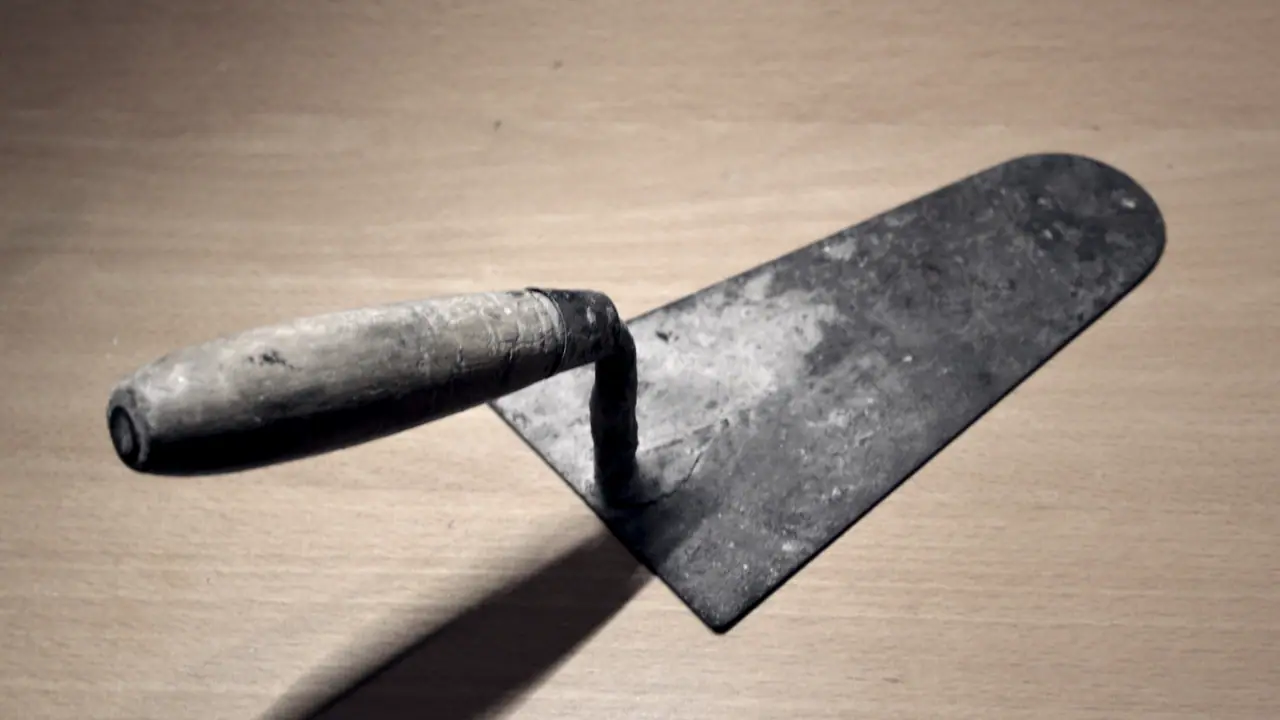 Truelle posée sur une table de bois, avec sa lame triangulaire usée, sa soie coudée et l'ombre qu'elle porte.
