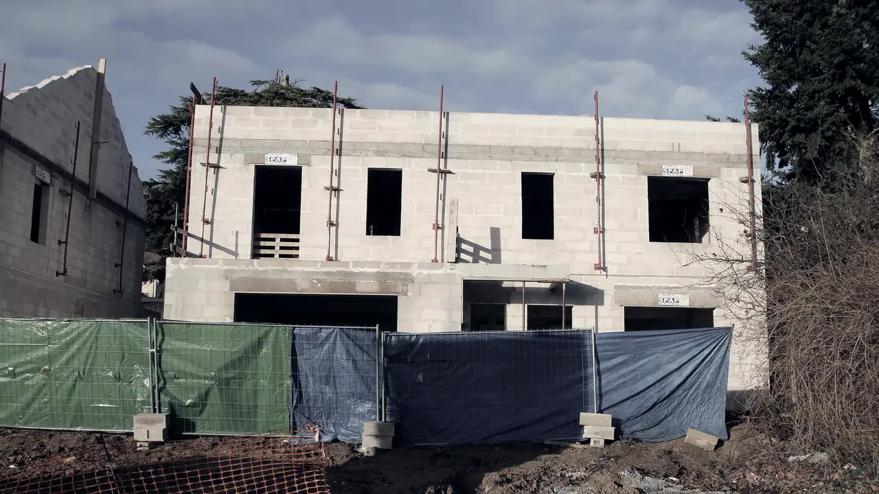 Maison en construction dont les murs sont montés, avec ses baies encore vides, ses étais obliques et la palissade de chantier devant elle.