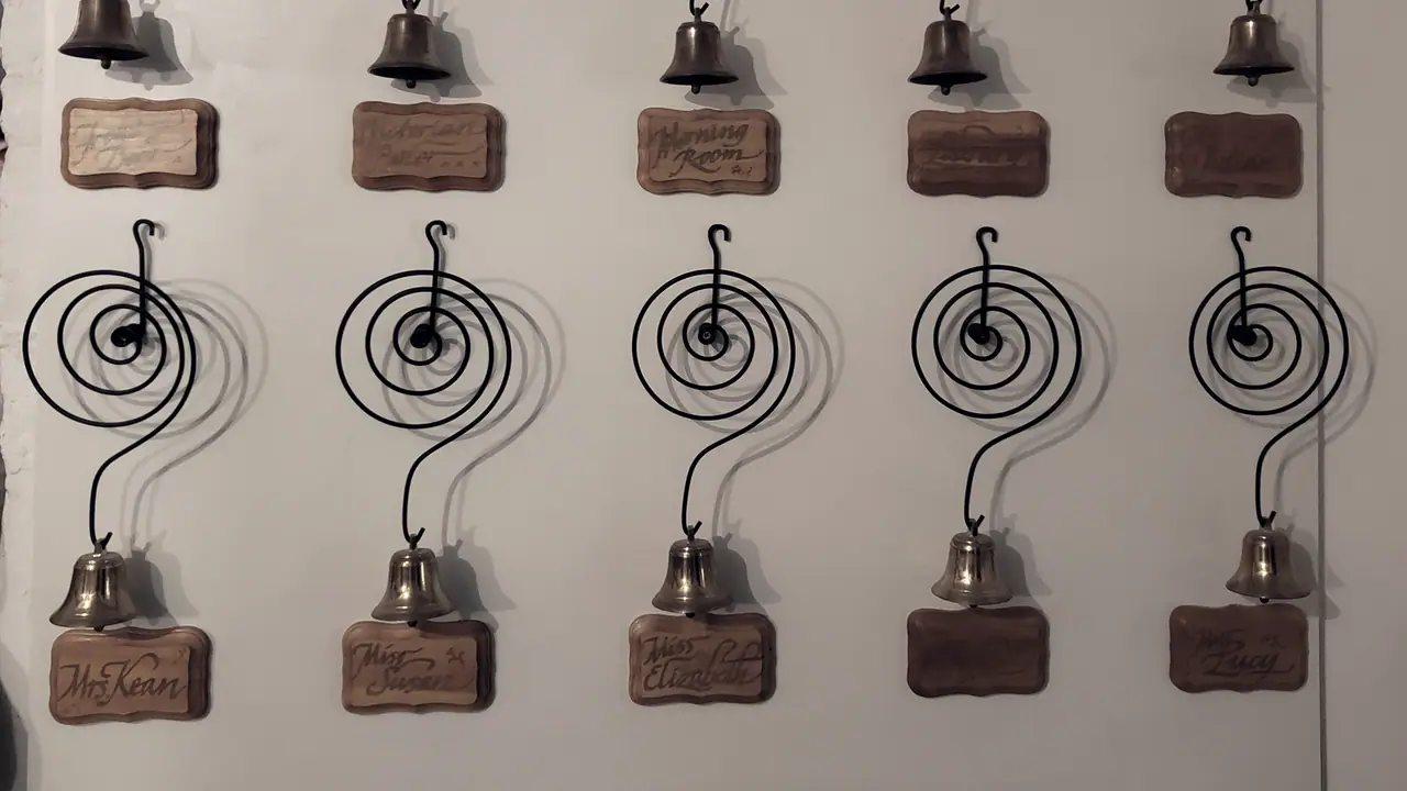 Clochettes de laiton suspendues à des ressorts en spirale sur un panneau clair, chacune au-dessus d'une plaquette de bois qui porte un nom.