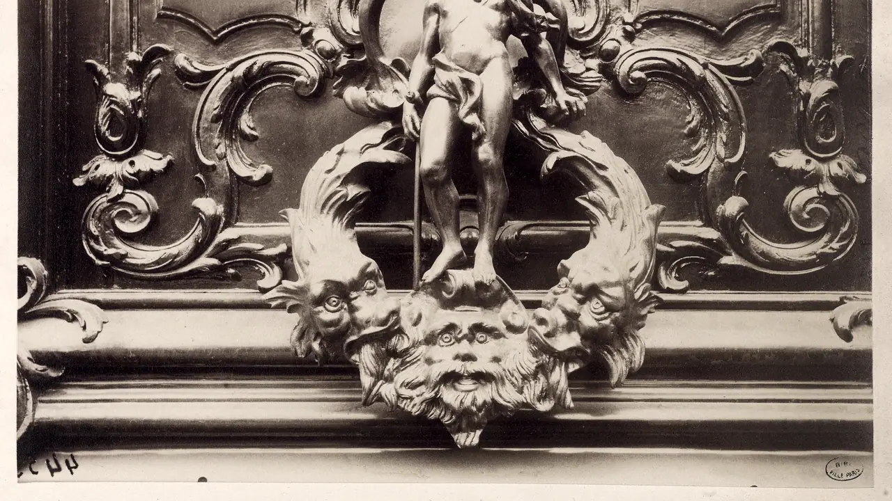 Photographie ancienne d'un heurtoir de bronze sur une porte sculptée : le bas d'une figure debout entre deux dauphins, au-dessus d'un masque barbu.