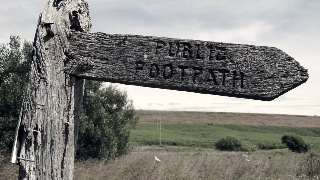 Vieux poteau indicateur de bois usé dont le bras taillé en flèche porte les mots Public footpath, devant un champ, des herbes sèches et quelques buissons.
