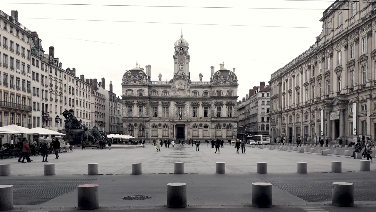 Grande place pavée devant un hôtel de ville à campanile, avec des passants entre les bornes de pierre au premier plan.