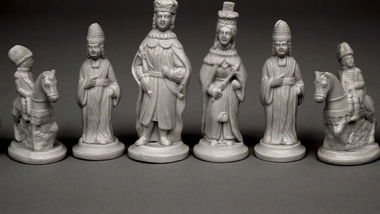 Six pièces d'échecs anciennes en céramique alignées devant un fond uni : deux cavaliers, deux fous coiffés d'une mitre, et au centre le roi et la reine.