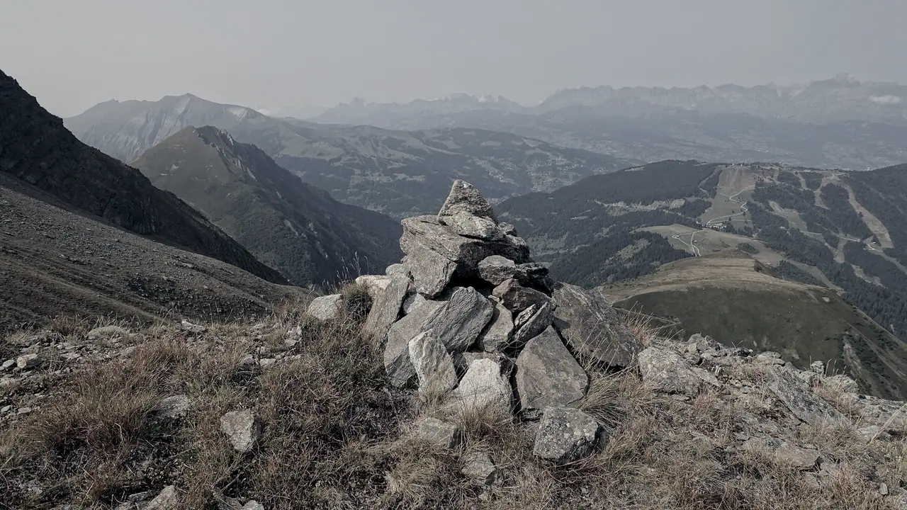 Cairn de pierres plates dressé sur une crête de montagne, devant des vallées boisées et des sommets qui s'estompent dans la brume.