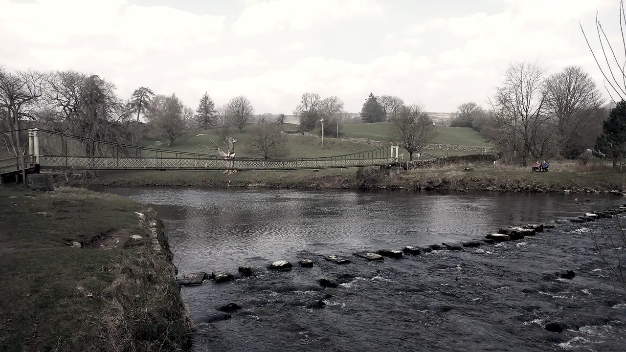 Pierres de gué alignées en travers d'une rivière, avec une passerelle suspendue qui franchit le même cours d'eau un peu plus loin.