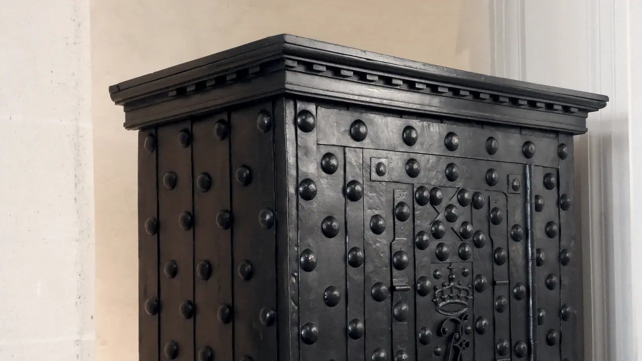 Coffre-fort ancien en fer clouté, avec un monogramme couronné forgé sur sa porte, posé contre un mur de pierre.