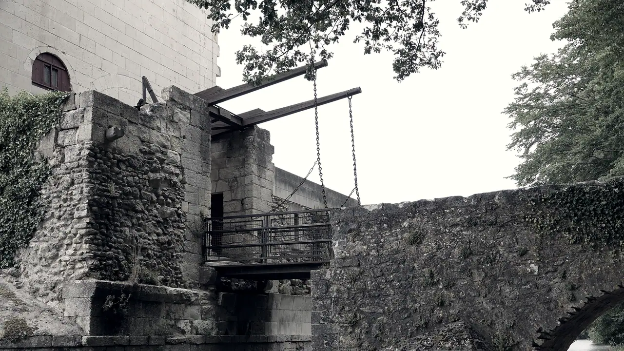 Pont-levis abaissé à l'entrée d'un château : deux flèches de bois et leurs chaînes tiennent un tablier à garde-corps grillagé, entre la porte et un vieux pont de pierre gagné par le lierre.