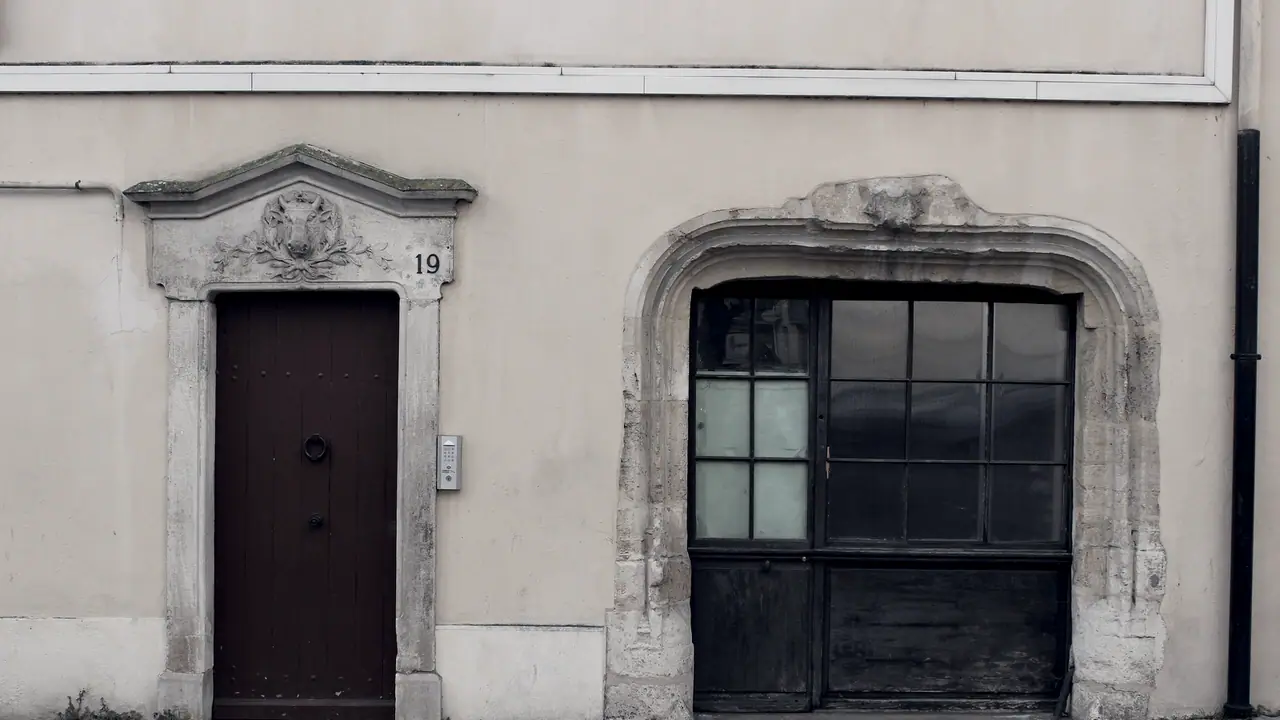Façade enduite percée de deux encadrements de pierre sculptée, une porte de bois à anneau sous un fronton portant le numéro 19, et une large baie cintrée fermée par un châssis vitré à petits carreaux.