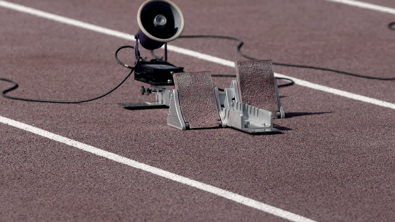 Blocs de départ posés sur une piste d'athlétisme entre deux lignes blanches, à côté du haut-parleur qui donnera le signal du départ.