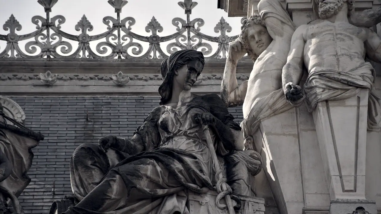 Statue allégorique assise au sommet d'une façade de palais, avec une balustrade en fonte ouvragée et deux atlantes sculptés.