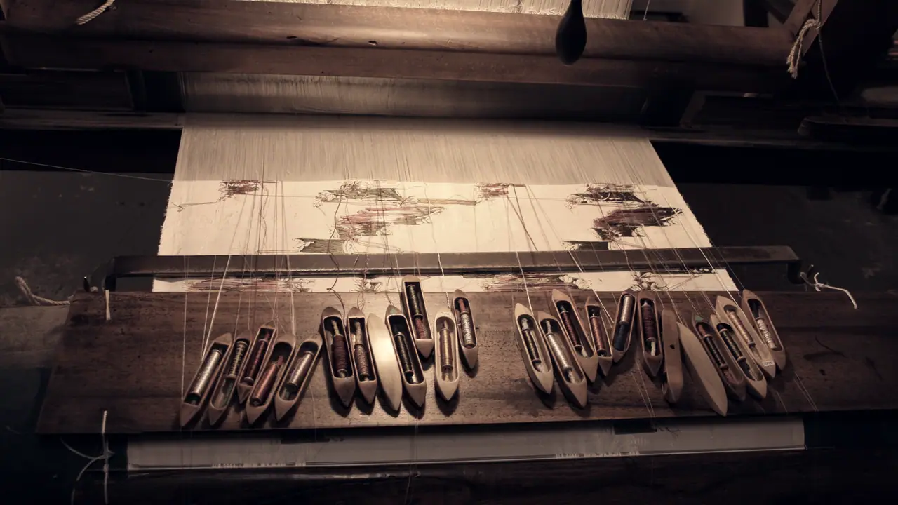 Navettes de bois alignées sur la planche d'un métier à tisser, leurs fils tendus vers une étoffe à motifs en cours de tissage.