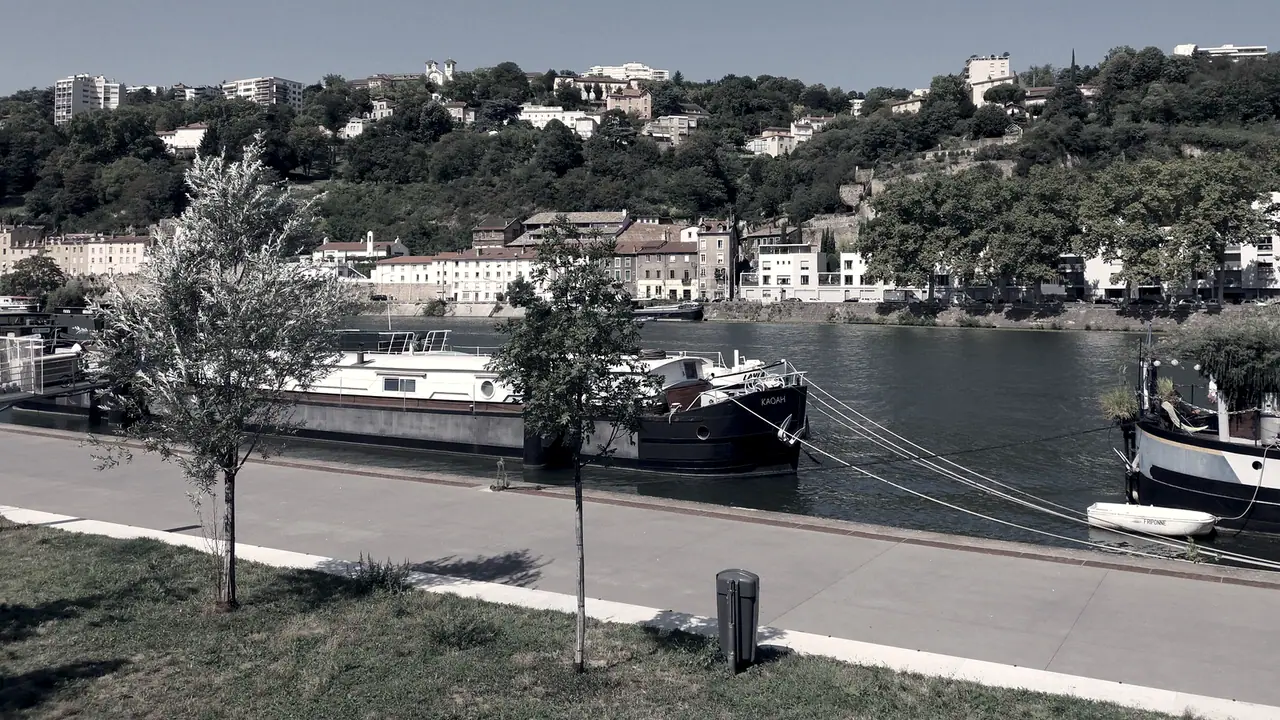 Péniches amarrées le long d'un quai en pierre, devant un coteau couvert de maisons et d'arbres au bord de la rivière.