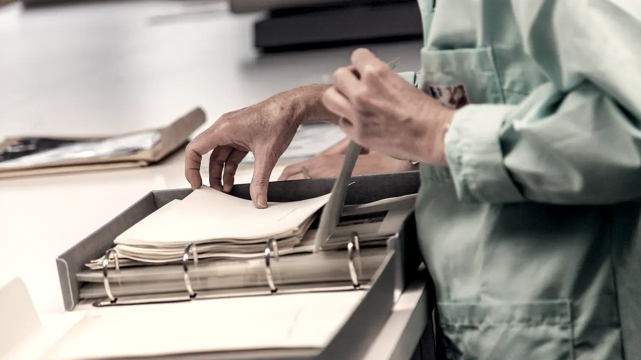 Mains d'une archiviste qui glissent des feuillets dans un classeur à anneaux posé dans sa boîte, sur une table de tri.