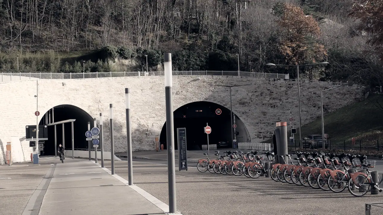Entrée d'un tunnel routier à deux voûtes dans un mur de pierre appareillée, surmontée d'un talus boisé, avec des vélos en libre-service rangés devant.