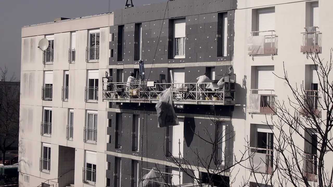 Deux ouvriers sur une nacelle suspendue devant une façade d'immeuble en partie recouverte de panneaux isolants.